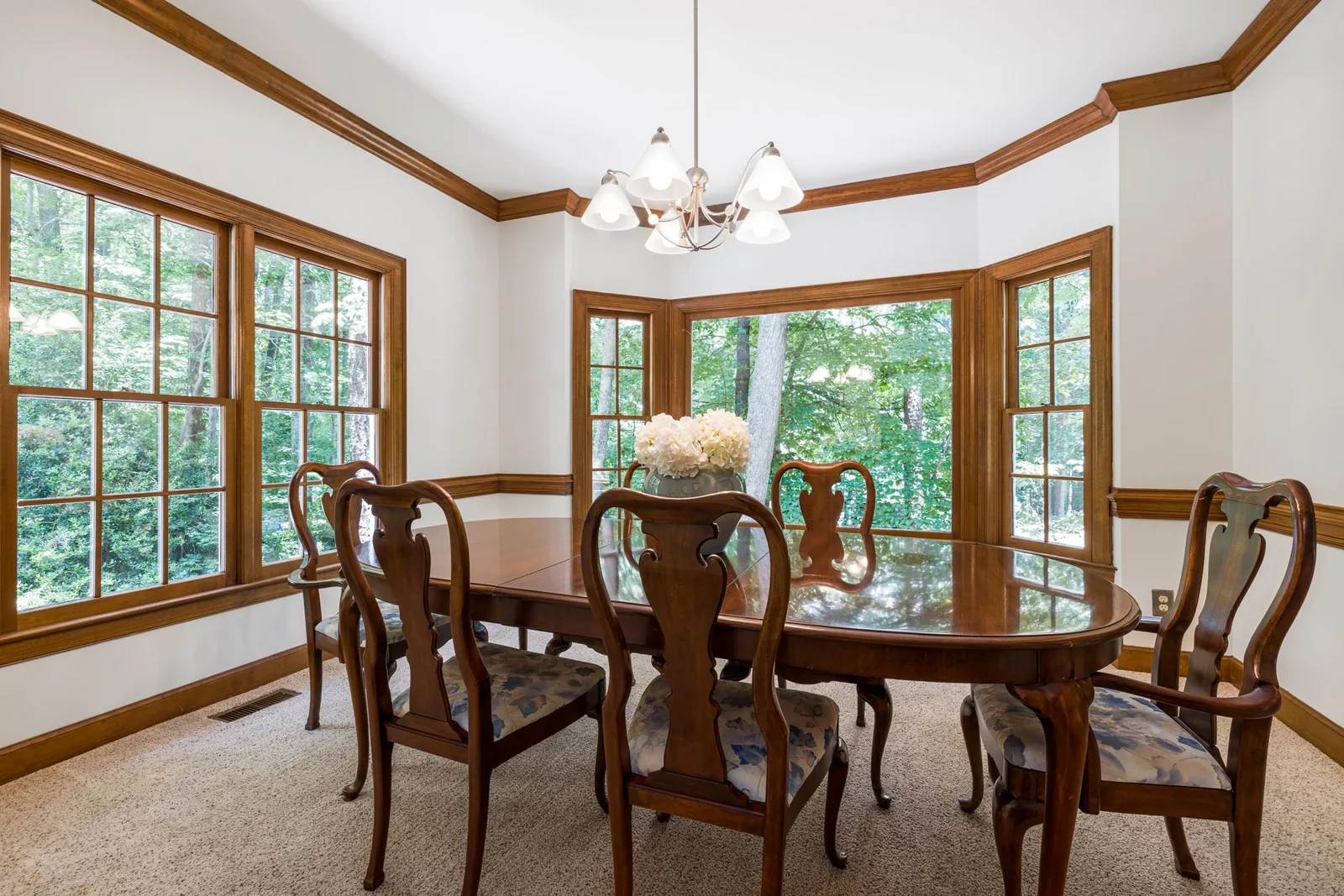Dining room with a wooden table and chairs beside large windows overlooking greenery.