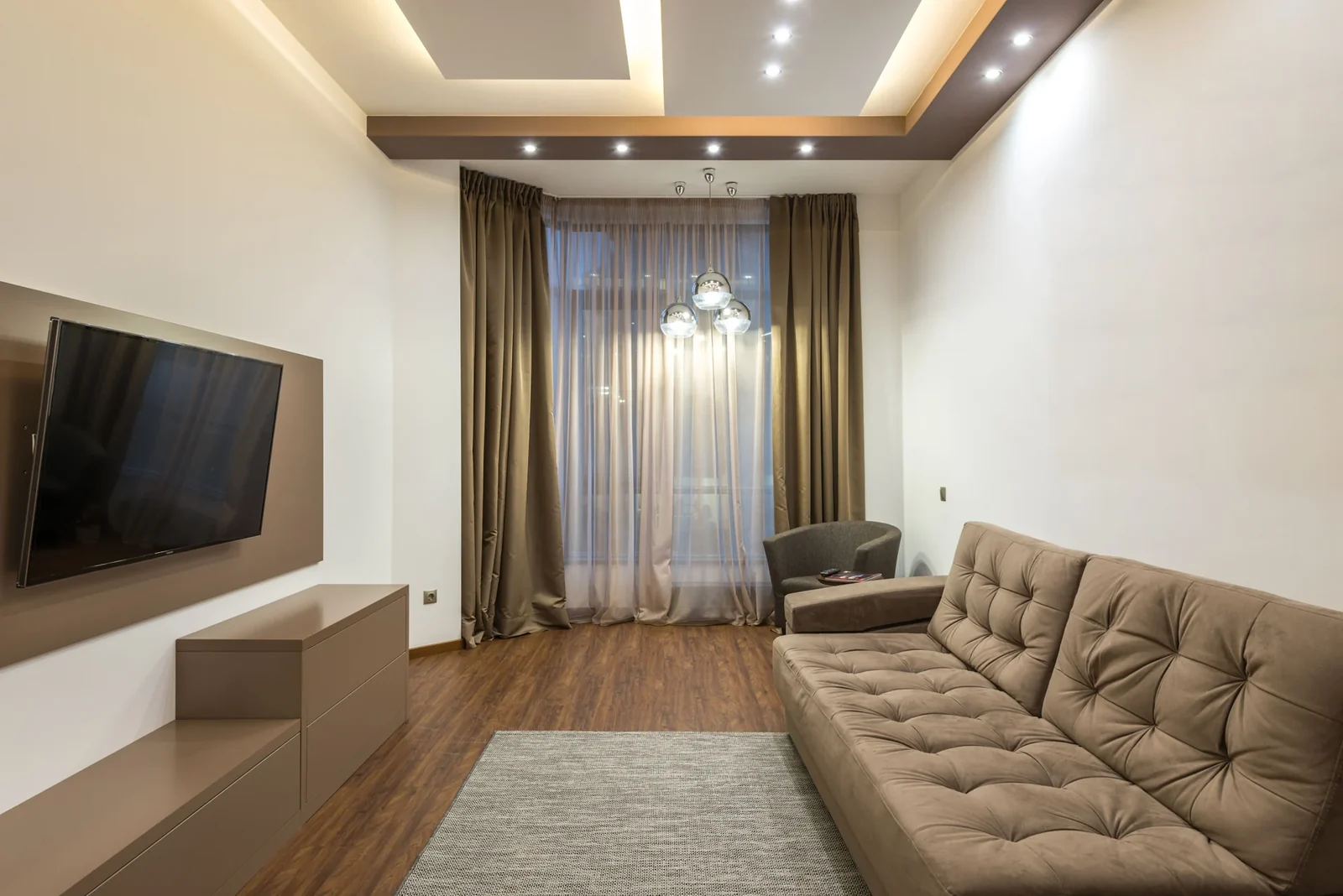 Beige living room with upholstered seating, an area rug and a wall-mounted television.
