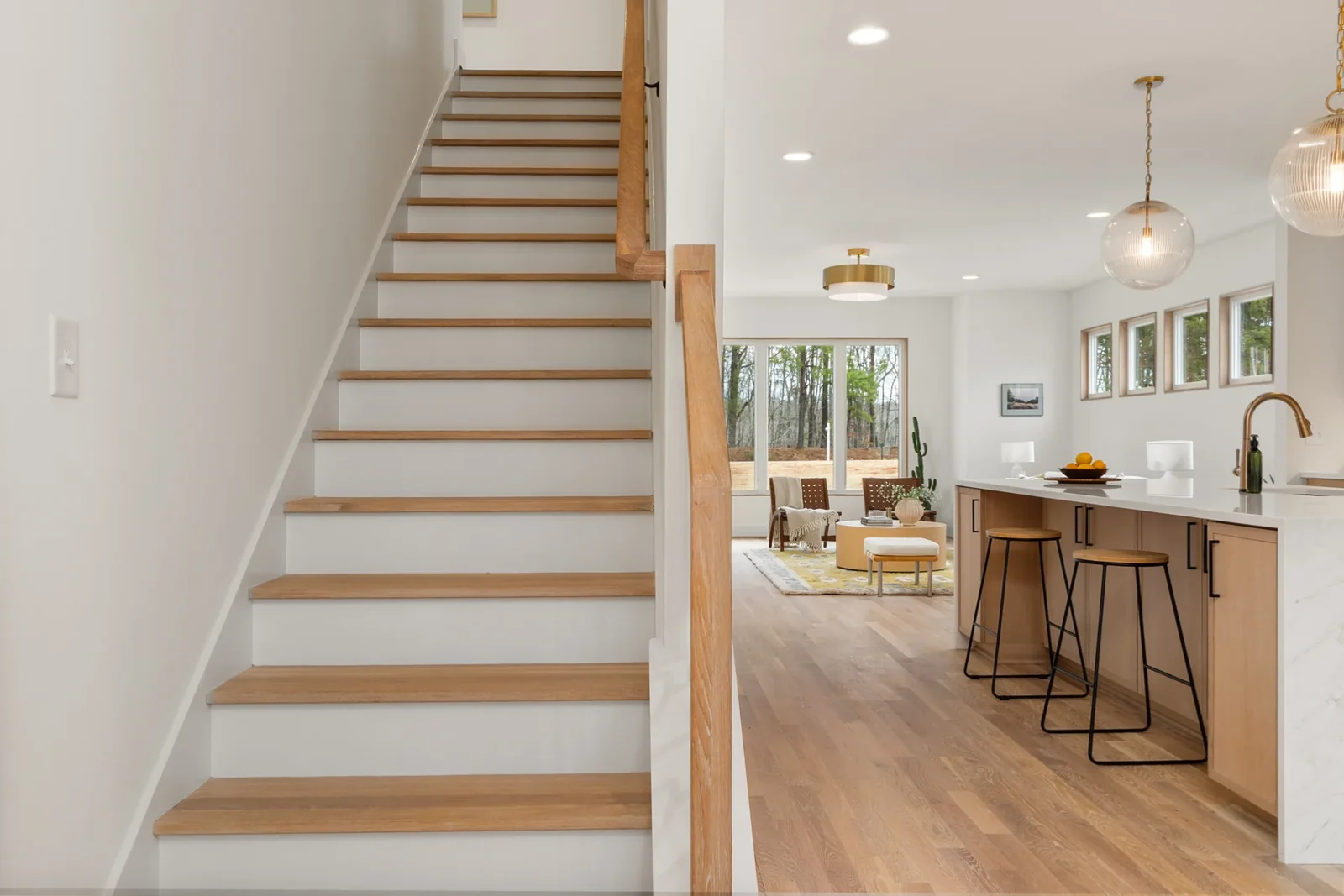 Bright open home interior with a wood staircase, oak kitchen island, and sitting area beside tall windows.