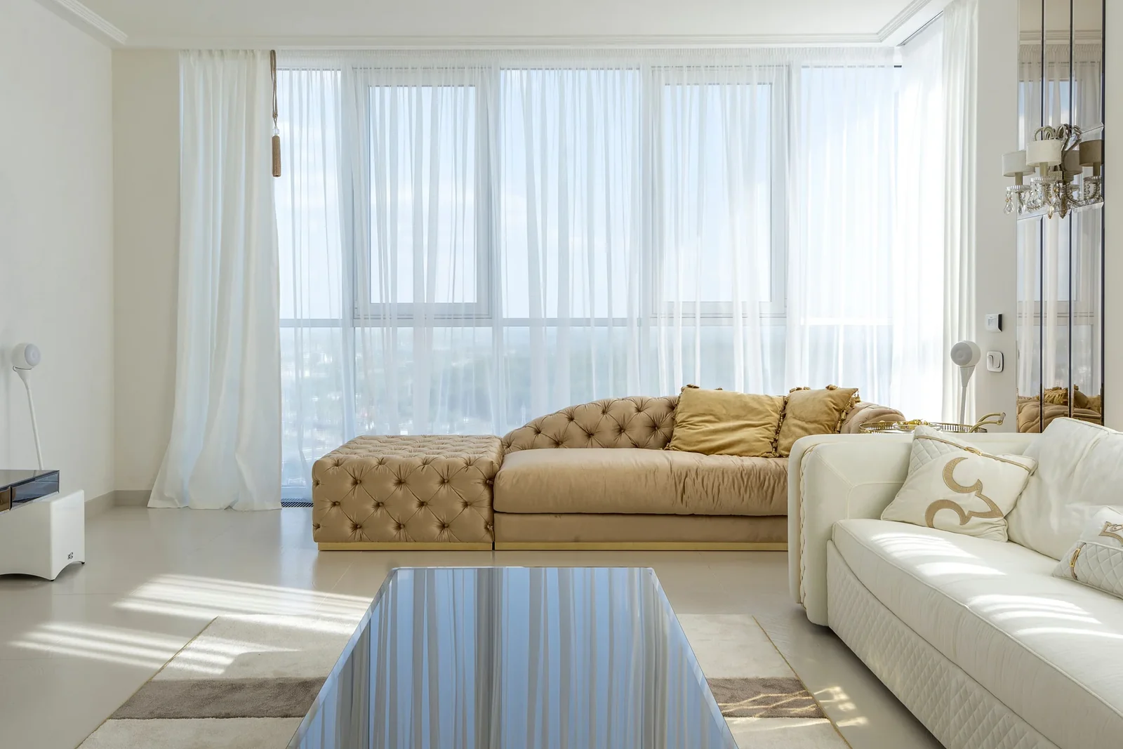 Light-filled living room with cream and tan sofas, sheer curtains, and a glass coffee table.