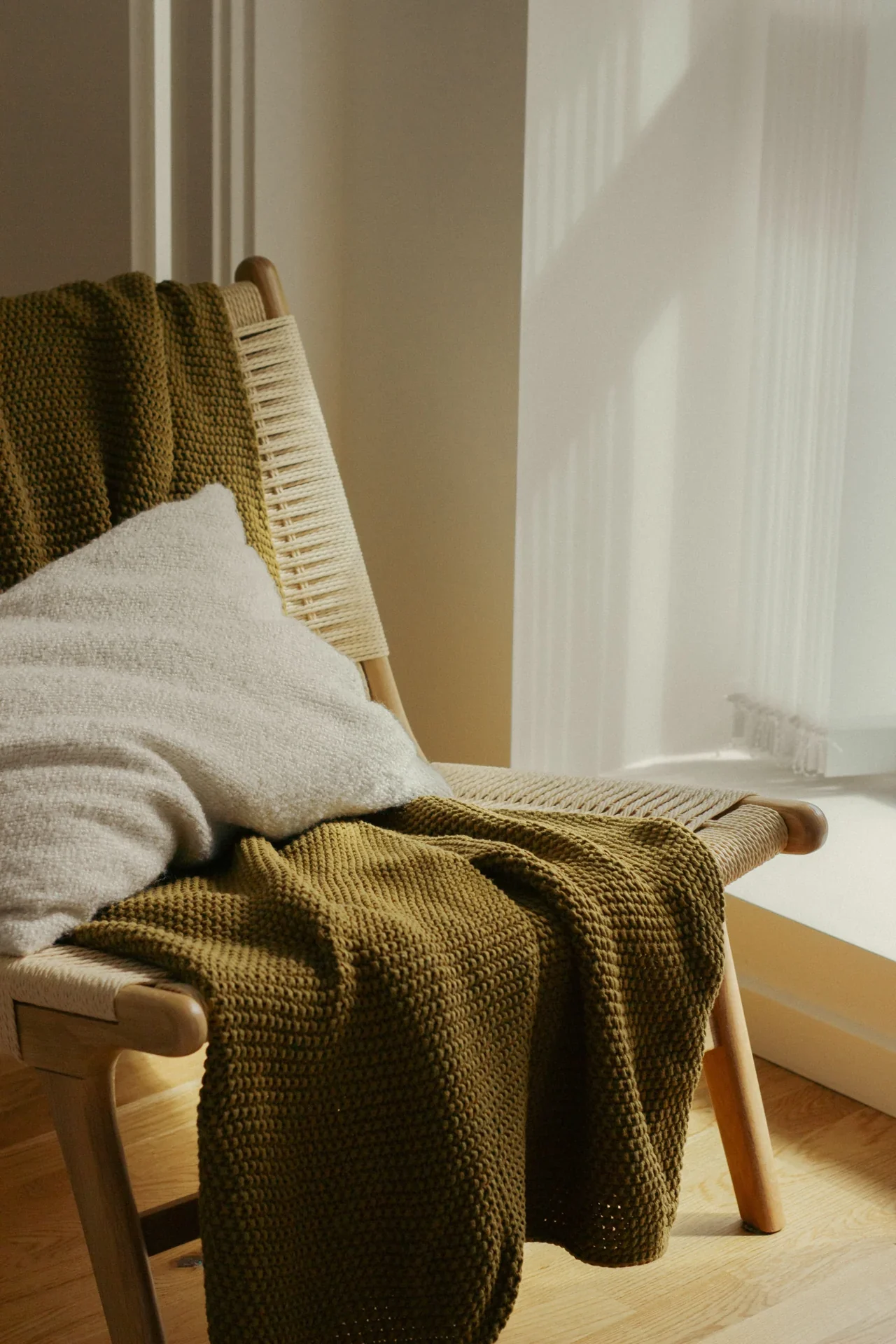 Chair with a green blanket and pillow beside a sunlit window.