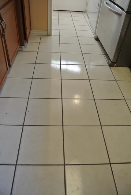 White square kitchen tile and grout before cleaning: Dark grout lines across a kitchen floor beside wood cabinets.