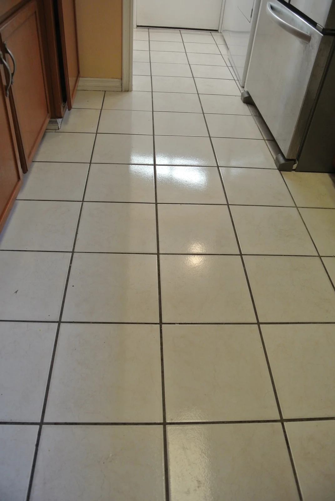 White square kitchen tile and grout before cleaning: Dark grout lines across a kitchen floor beside wood cabinets.